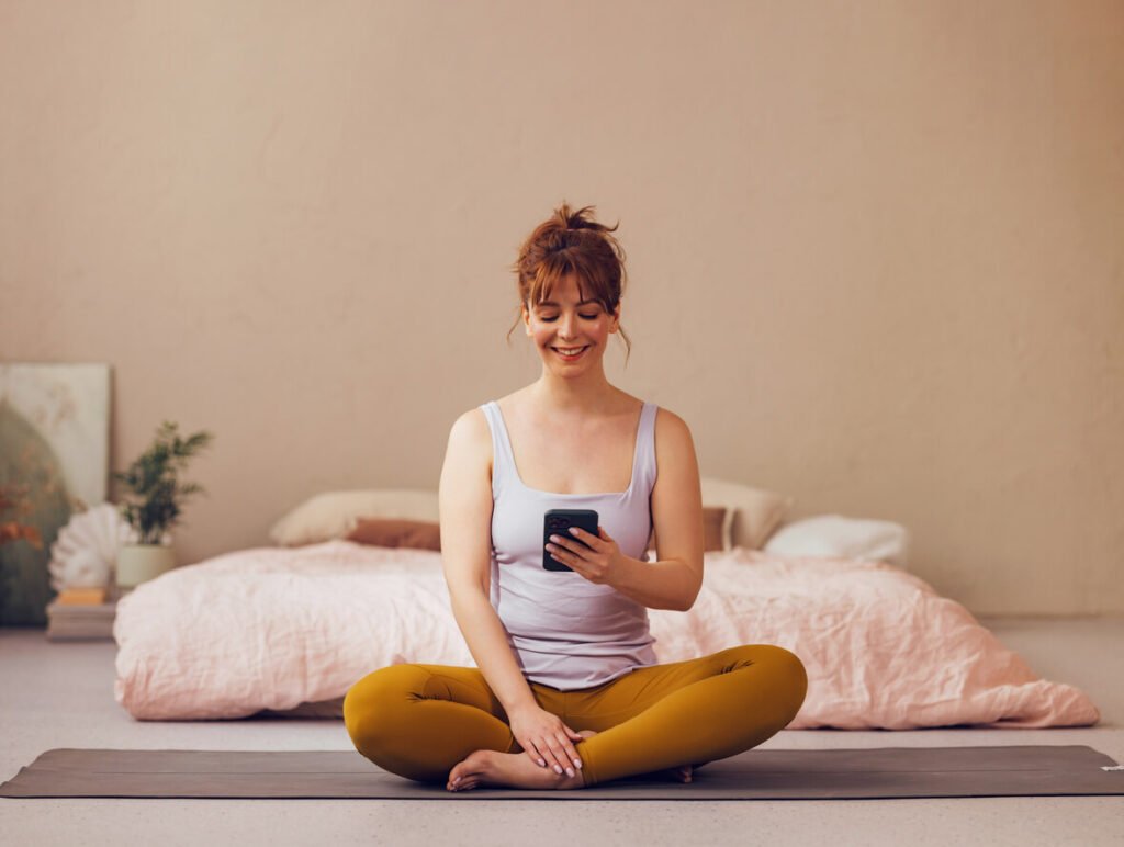 Smiling Woman Using Smartphone on Yoga Mat in Cozy Bedroom stock photo. jenniferanistonbody.com