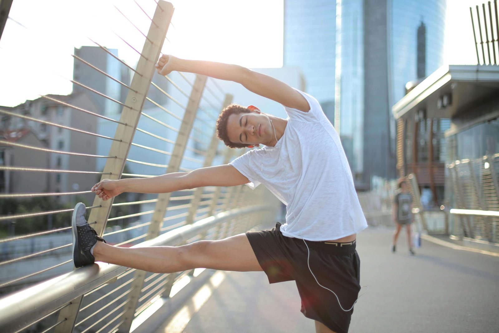 Young slender ethnic sportsman in earbuds warming up and listening to music with pleasure on street in city. jenniferanistonbody.com